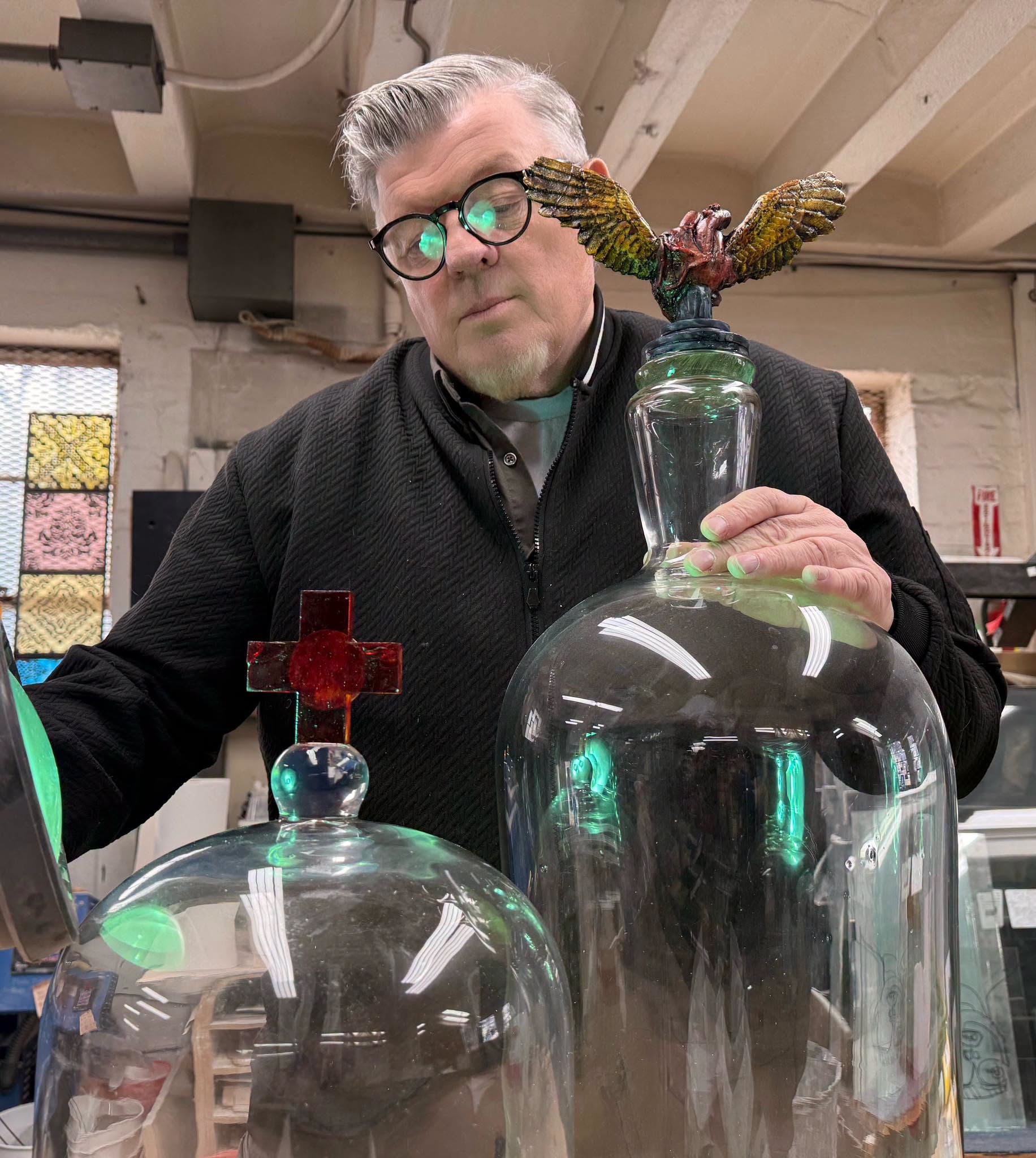 Portrait of the artist Tim Tate in his studio, holding a blown glass dome topped with a winged sacred heart